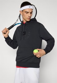 Black hoodie with a logo print, featuring a front pocket, paired with white shorts and a red undershirt. Holding a tennis ball and racket.