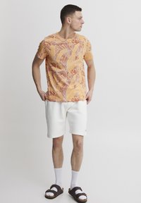 Camisa de hombre de manga corta con un estampado naranja que presenta un diseño de hojas, combinada con pantalones cortos blancos, calcetines blancos y sandalias marrones.