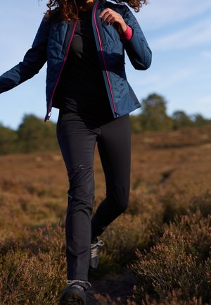 Veste isolante bleue avec des accents roses, pantalon noir ajusté et chaussures de randonnée noires. Décor extérieur avec de la végétation de bruyère.