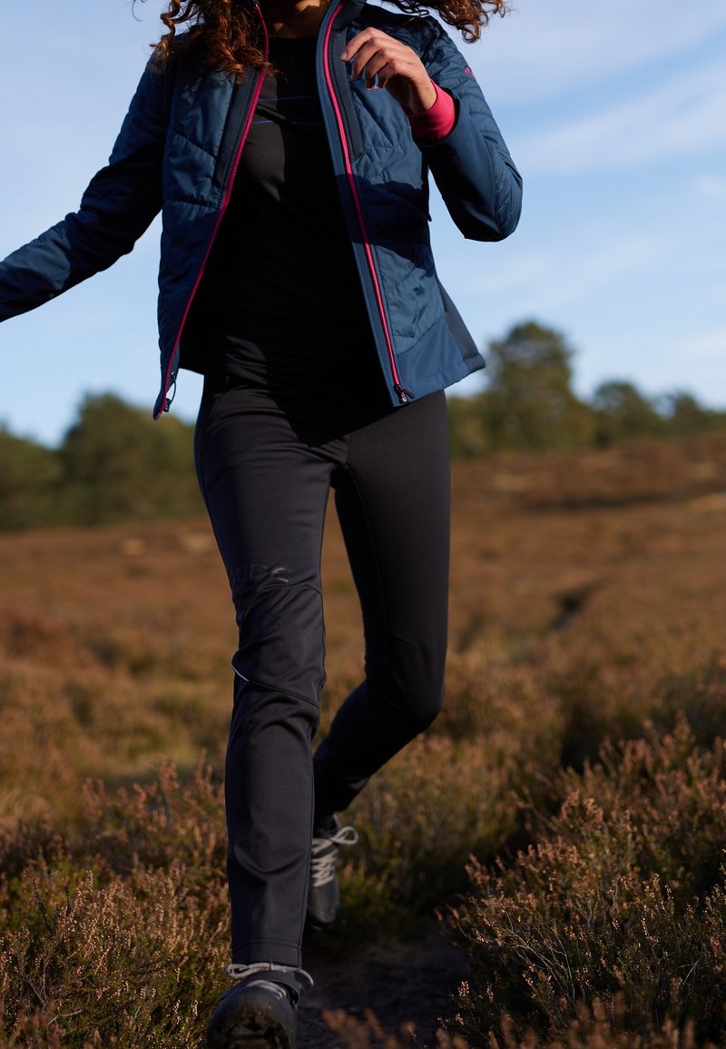 Blå isolerad jacka med rosa detaljer, svarta tajta byxor och svarta vandringsskor. Utomhusmiljö med ljungvegetation.