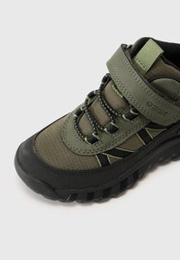 Chaussure mi-haute en tissu vert et caoutchouc avec accents noirs, dotée d'une sangle en Velcro et d'une semelle texturée, mise en valeur par des coutures vertes.