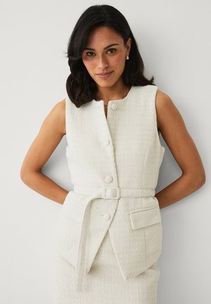 Frau mit dunklen Haaren, die eine ärmellose weiße strukturierte Weste mit Knöpfen, Gürtel und Taschen trägt und vor einem einfarbigen hellen Hintergrund posiert.