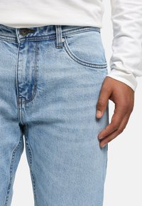 Jeans de mezclilla azul claro con corte relajado, que presentan cinco bolsillos, costuras beige y un cierre de botón clásico en la cintura.