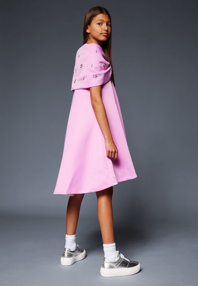 Ružičasta haljina do koljena s napuhanim rukavima i cvjetnim ukrasima. Uparena s metalik srebrnim platformskim tenisicama i bijelim čarapama.