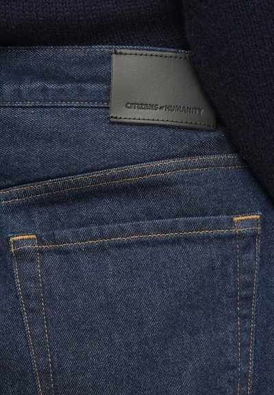 Sötétkék farmernadrág narancssárga varrással, fekete bőr címkével a hátoldali zseb közelében, amelyre a "CITIZENS of HUMANITY" felirat van nyomva.
