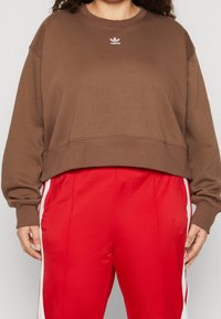 Person som bär en brun kortärmad Adidas-tröja och röda träningsbyxor med vita sidostreck, står med händerna avspänt vid sidorna.