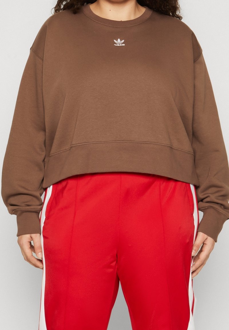 Person som bär en brun kortärmad Adidas-tröja och röda träningsbyxor med vita sidostreck, står med händerna avspänt vid sidorna.