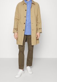 Beżowy płaszcz trencz zapinany na guziki, noszony na jasnoniebieskiej koszuli, w połączeniu z oliwkowozielonymi spodniami i białymi trampkami.