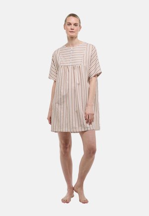 Femme debout, pieds nus, portant une robe ample mi-longue aux rayures verticales beige et blanches avec des manches courtes.