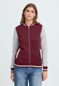 Sweat-shirt zippé bordeaux avec manches gris clair, deux poches avant, poignets et col côtelés. Fabriqué en matériau doux. Présente une étiquette logo.