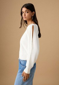 Bel pulover iz pletenine z odprtimi rameni in rebrastimi manšetami, kombiniran s svetlo modrimi kavbojkami. Tkanina z teksturo, sproščen kroj.