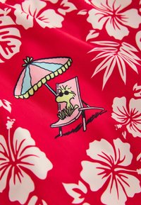 Tissu rouge avec des fleurs blanches d’hibiscus et un oiseau de dessin animé portant des lunettes de soleil assis sur une chaise longue sous un parasol rayé.