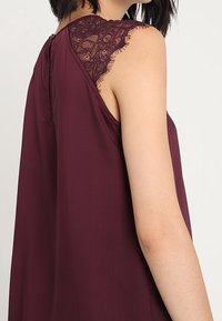 Top burdeos con detalles de encaje en los hombros, diseño fluido y escote en forma de V en la parte posterior. La tela parece suave y ligera.