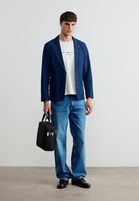 Νεαρός άνδρας που φοράει μπλε σκούρο σακάκι, λευκό μπλουζάκι με γραφικά, μπλε τζιν, μαύρα παπούτσια, κρατώντας μαύρη τσάντα χειρός, στέκεται μπροστά σε απλό λευκό τοίχο.