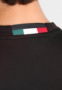 Musta kangaspaita, jossa on teksturoitu kaulus. Selässä on pieni logo, jossa on Italian lipun värit: vihreä, valkoinen ja punainen.