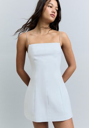 Junge Frau mit langen dunklen Haaren, die ein ärmelloses, eng anliegendes weißes Minikleid mit dünnen Trägern vor einem schlichten hellen Hintergrund trägt.