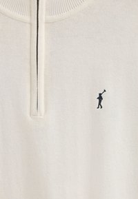 Krämfärgad tröja med halvdragkedja vid kragen. Har ribbad textur och en liten svart silhuettlogotyp av en person med en golfklubba.