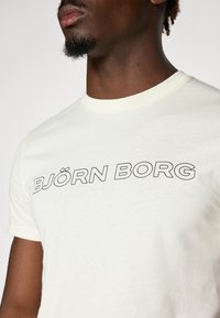 Helles Cremefarbenes T-Shirt mit kurzen Ärmeln, das auf der Vorderseite in fettem Schwarz den Schriftzug "BJÖRN BORG" trägt. Hergestellt aus weichem, glattem Stoff mit einem klassischen Rundhalsausschnitt.