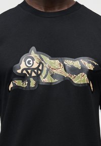 Μαύρο βαμβακερό μπλουζάκι με στυλιζαρισμένο γραφικό ενός πλάσματος με καμουφλάζ μοτίβο, περιγραμμένο με γκρι, με μεγάλα μάτια και αιχμηρά δόντια.