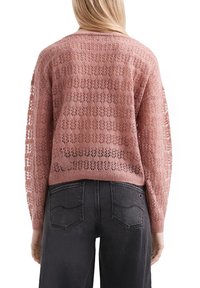 Frau, die einen transparenten rosa Strickpulli mit floralen Mustern und dunklen grauen Jeans trägt, von hinten gezeigt.