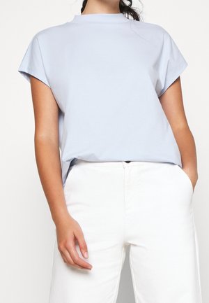 Kobieta w jasnoniebieskiej bluzce z krótkim rękawem, włożonej do białych spodni z wysokim stanem, z jedną ręką w kieszeni.