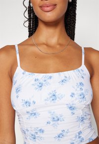 Top sin mangas blanco con flores y rosas azules, cuenta con tirantes finos y laterales arrugados para un ajuste ceñido. Textura de tejido de algodón suave.