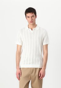 Camisa polo branca de manga curta com um padrão de malha texturizada listrada e um design em V, emparelhada com calças bege.