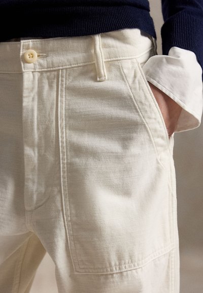 Persoană purtând pantaloni de culoare crem deschis, cu mâna în buzunar, pulover bleumarin și manșetă albă de cămașă vizibilă la încheietură.