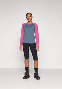 Sporttop met lange mouwen, grijze body en roze mouwen, gecombineerd met zwarte capri-legging en zwarte schoenen. Belangrijk merkdetail op de top.