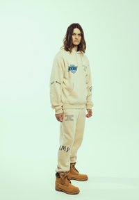 Sudadera beige con gráfico azul, texto en las mangas y pantalones de chándal a juego. Botas beige con diseño de cordones. Textura de tela suave.