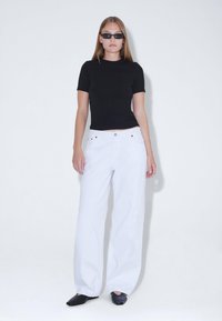 Top negro ajustado de manga corta, pantalones anchos blancos con detalles de botones y zapatos planos negros. La modelo posa frente a un fondo blanco.