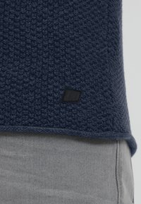 Marinefarget teksturert genserstoff med et vevet mønster, med et svart logo-lapp på nederkanten. Under er lys grå stoff fra bukser synlig.