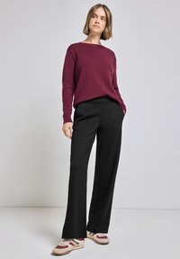 Bordeaux gebreide trui gecombineerd met losse zwarte broeken. Model draagt beige en bordeaux sneakers. Zachte texturen, casual pasvorm en recht toelopende broekspijpen.