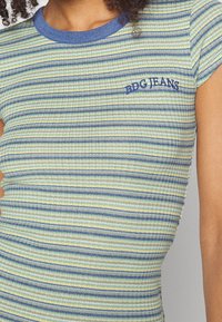 Žebrovaný krátký rukávový top s proužky v modré, zelené a krémové barvě; na hrudi je vyšívaný nápis "BDG JEANS" modrou barvou.