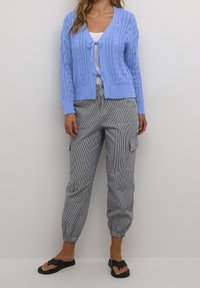 Jasnoniebieski dziergany sweter na guziki z wzorami warkoczy, z otwartym przodem i wiązaniem, zestawiony z pasiastymi spodniami cargo i czarnymi klapkami.