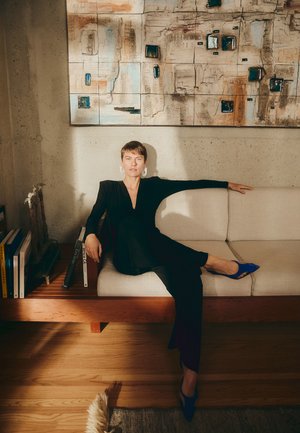 Vestido negro de manga larga con escote en V profundo, tacones azules de punta, sentada en un sofá beige claro con acentos de madera, contra una pared texturizada.