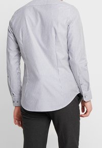 Randig långärmad skjorta i blått och vitt. Har knäppning vid ärmsluten, en rundad nederkant och en mjuk bomullstextil. Bakifrån syns sömdetaljer.