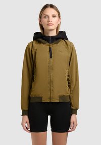 Junge Frau trägt eine olivgrüne Zip-up-Jacke mit schwarzer Kapuze und schwarzen Radlershorts, steht vor einem einfarbigen Hintergrund.