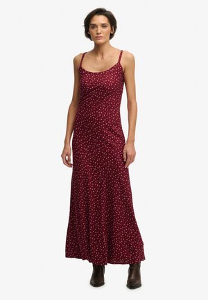 Kobieta ubrana w bezrękawnikową, bordową sukienkę maxi z małym kwiatowym wzorem i brązowe botki, stojąca na białym tle.