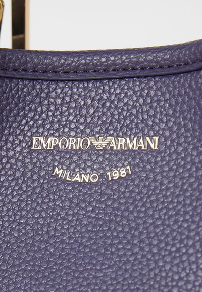 Prim-plan al unei suprafețe din piele texturată de culoare violet, cu logoul embosat "EMPORIO ARMANI" și "MILANO 1981" în litere aurii.