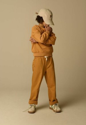 Enfant portant un sweat-shirt beige, un pantalon assorti et une casquette beige, debout les bras croisés et la main sur le menton dans une pose contemplative sur un fond beige uni.