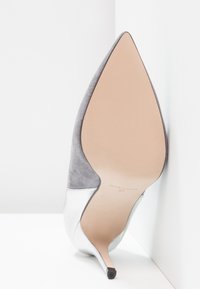 Chaussure en daim gris à bout pointu avec un accent métallique argenté, talon stiletto fin et semelle beige lisse, positionnée sur un fond blanc.
