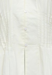 Vêtement blanc boutonné avec des plis verticaux sur les côtés et un pli central devant qui descend vers le bas.