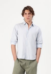 Camisa de botones a rayas azul claro hecha de tela suave, con cuello clásico, mangas enrolladas y un ajuste relajado. Combinada con pantalones verdes oliva.