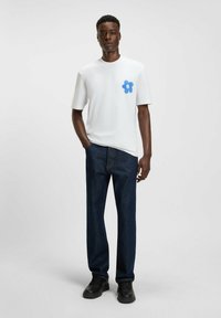 White short-sleeve t-shirt featuring a blue flower graphic on the left chest, paired with dark blue jeans and black shoes.