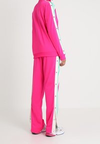 Persona vestida con un chándal rosa brillante con botones a presión en rayas blancas y azul claro en las mangas y las piernas del pantalón, de pie de lado.