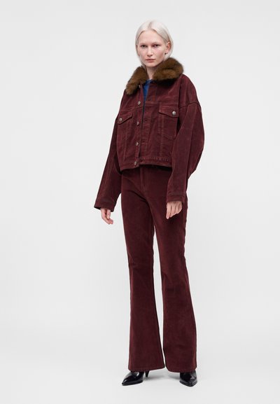 Femme aux cheveux pâles portant une veste en velours côtelé bordeaux avec un col en fourrure marron et un pantalon évasé assorti avec des chaussures à talons noires.