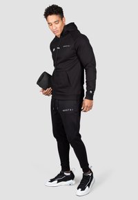 Set athletic negru, format dintr-un hanorac cu glugă, dotat cu un buzunar tip kangur și un logo, asortat cu pantaloni jogger și pantofi sport negri cu alb.