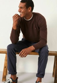 Brauner Strickpullover mit gerippten Bündchen, kombiniert mit marineblauen Hosen. Der Mann sitzt auf einer Holzbank, lächelt und trägt weiße und blaue Sneaker.