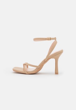 Nøgen højhælet sandal med ankelrem, lavet af et glat syntetisk materiale, firkantet tådesign og tynde remme, der krydser over foden.